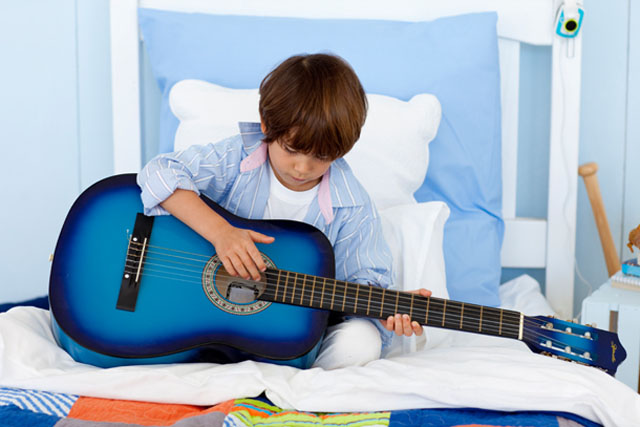CHAMBRES_ENFANTS_Photo_640X427_Garçon_assis_sur_son_lit_et_jouant_de_la_guitare CHAMBRES_ENFANTS_Photo_640X427-Jeune_garçon_confortablement_assis_sur_son_lit_pour_jouer_de_la_guitare_bien_adossé_à_des_oreillers_et_un_gros_coussin